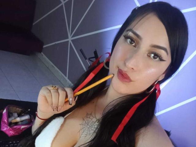 Chloey69, avec de longs cheveux noirs, des rubans rouges, tenant un crayon jaune, regardant la caméra, portant un haut blanc, avec un tatouage de toile d'araignée visible, du rouge à lèvres, des boucles d'oreilles cerceaux, et un sac rose en arrière-plan..