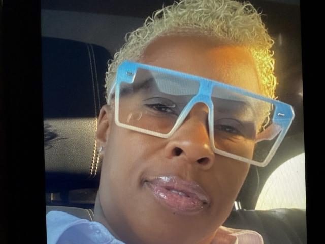 PinkyX69 tiene la piel de color marrón claro, cabello rubio rizado y lleva unas gafas azules grandes con labios rosados brillantes. Está sentada en un coche con un asiento de cuero negro..