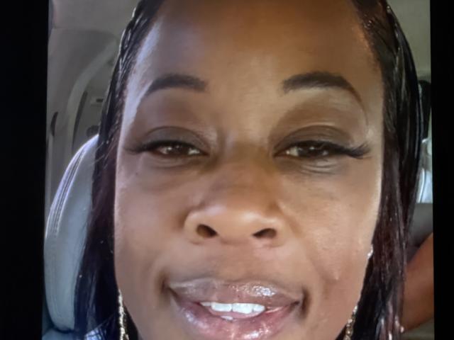 Close-up of PinkyX69's face, smiling slightly. She has dark brown skin, glossy lips with a hint of gloss, and long black braids. Her eyebrows are dark and well-defined. She's wearing small gold earrings. Her eyes are half-open, with long eyelashes. The image has a natural light source, highlighting her skin's texture. The background is blurred, focusing on her face. The photo is realistic, with a slight soft focus on her lips.