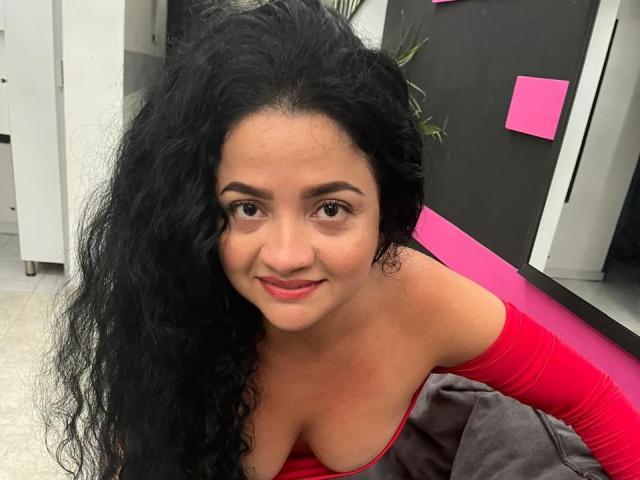 LinaSuart with long black curly hair, red off-shoulder top, showing her large, bare, round, perky tits, smiling at the camera, sitting on a gray chair.