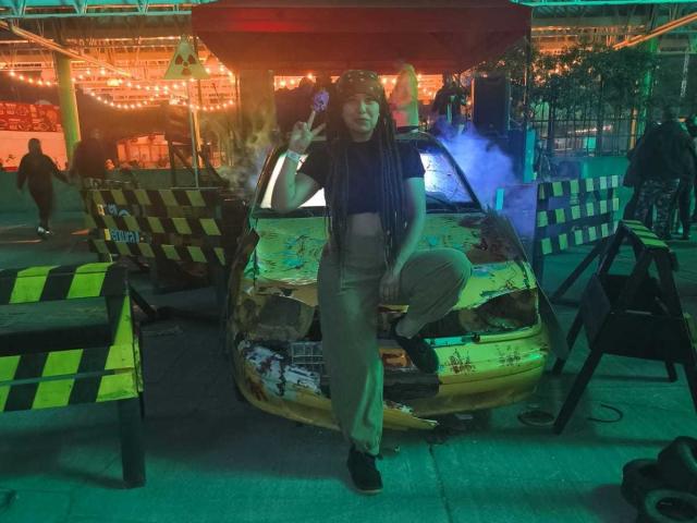 BeckyHuston stands in front of a heavily damaged yellow car with graffiti and broken windows. She's wearing a dark blue top, beige pants, and black shoes. She's making a peace sign with her right hand. The car is surrounded by black and yellow striped barriers. The scene is illuminated by green and orange lights, with smoke rising from the car. The background is blurry, with people and string lights. The image has a gritty, urban vibe.