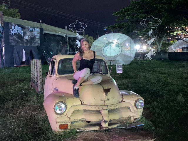 BrigitteWalsh with tits in a black top, sitting on a yellow, rusty vintage car at night, wearing white pants and black boots, with a large umbrella behind her.