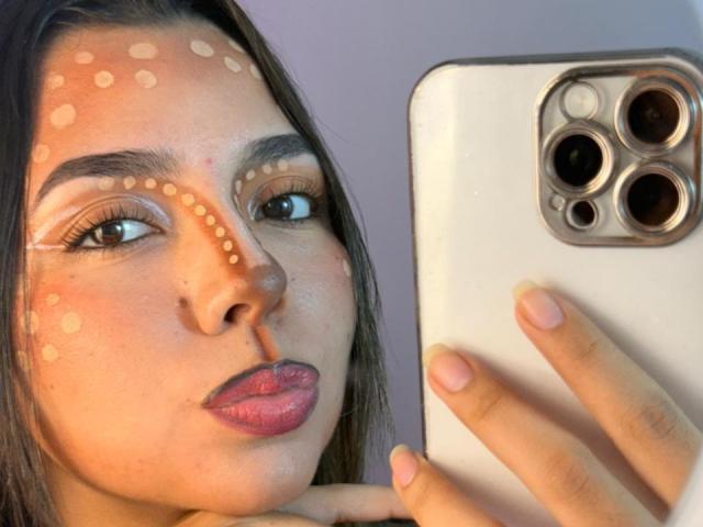 Das Gesicht von LatinaIu mit weißen Punktmustern auf den Wangen und der Stirn, die ein weißes iPhone mit drei Kameras hält, mit rosa Lippenstift, langen braunen Haaren, rosa Nägeln und einem neutralen Gesichtsausdruck..