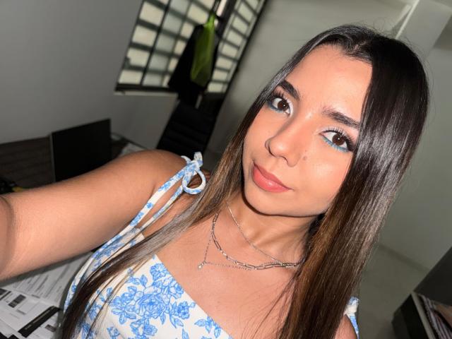 LatinaIu mit langen, braunen Haaren, blauem Augen-Make-up und einem Nasenring, trägt ein blaues, geblümtes Kleid, lächelt in die Kamera, in einem gut beleuchteten Raum, und zeigt ihre Brüste..
