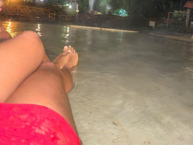 Le gambe di LatinaIu in un abito rosso, seduta in acqua bassa, che mostra i suoi piedi nudi con un leggero riflesso, di notte, con uno sfondo sfocato..