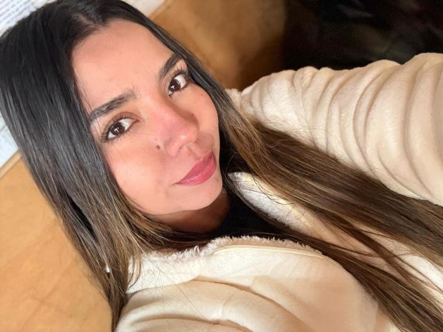 LatinaIu, con lunghi capelli castani, occhi castani, rossetto rosa chiaro e un sorriso carino, indossa una giacca beige con zip, guarda verso la fotocamera, in un selfie..