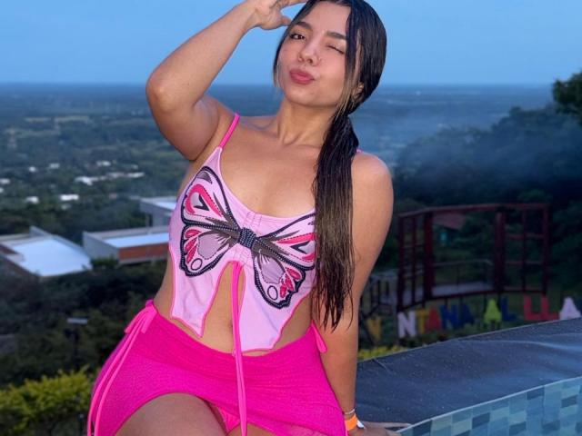 LatinaIu with long brown ponytail, pink butterfly top showing her tits, and pink shorts, sitting by a pool, winking, and touching her forehead. Blue sky in the background.