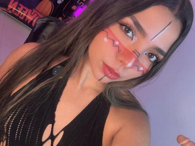 LatinaIu con lunghi capelli castani, una maglietta di rete nera e dello sperma sul viso e sulla fronte. Ha il trucco rosso, l'eyeliner nero e un naso piccolo. Le sue labbra sono leggermente socchiuse..