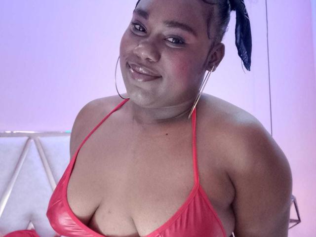 SweetyChocolatte, a plus-sized woman with dark skin, wears a red, shiny, halter-style bikini top that reveals her large cleavage. She has a black, woven hair accessory and wears large hoop earrings. She smiles softly, showing a slight blush on her cheeks. The light in the room has a purple-blue tint, highlighting her skin and the shiny texture of her top. Her relaxed posture and confident expression dominate the composition.