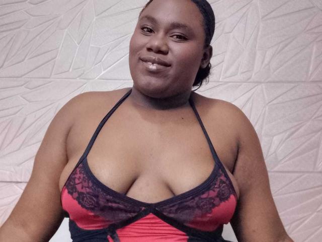 SweetyChocolatte, a dark-skinned woman with a confident smile, is centered in the image. She has a full, curvy figure and is wearing a red and black lace halter top that highlights her large breasts. Her hair is pulled back, and she has a subtle sheen on her face. The textured, geometric background is white and contrasts with her dark skin. The image is a photograph.
