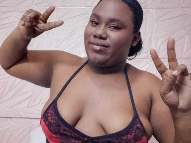 SweetyChocolatte with dark skin, black lace halter top, showing off her big, round, shiny tits, making rock-and-roll hands, smiling confidently, and having a cute, sexy, attractive look.