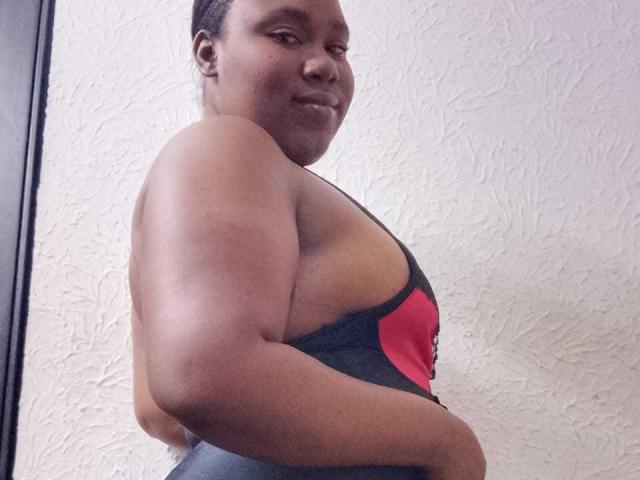 SweetyChocolatte, con piel morena, cabello corto trenzado y una sonrisa segura, vestida con un top deportivo negro y rojo, mostrando brazos fuertes y redondeados, y un trasero curvilíneo y voluminoso..