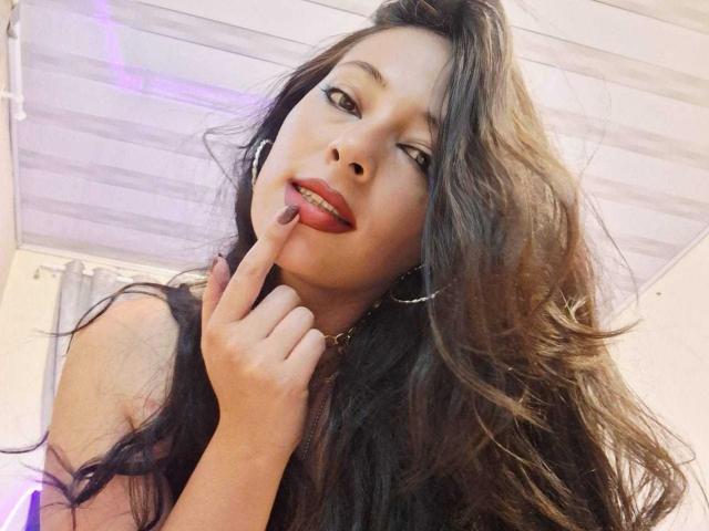 JessyLuxs with long brown wavy hair, red lipstick, and dark eyes, touching her red lip with her finger, wearing hoop earrings, smiling slightly, in a selfie.