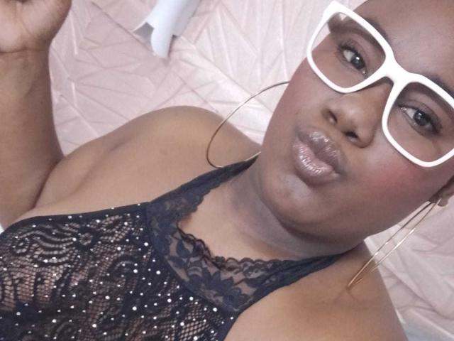 SweetyChocolatte with big white glasses, dark skin, pouty lips, and a lace black halter top showing her boobs. She's got gold hoop earrings and a confident smile.