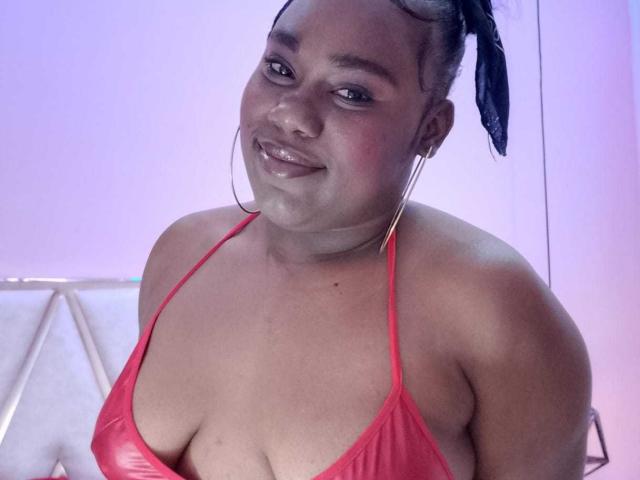 SweetyChocolatte, con piel morena, cabello trenzado, aros grandes y un top rojo de tirantes que muestra sus grandes y brillantes senos. Está sonriendo y mirando a la cámara..