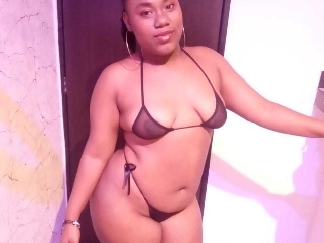 SweetyChocolatte trägt einen winzigen schwarzen Bikini aus Netzmaterial, der ihre kleinen Brüste, ihren runden Hintern und ihre dunkle Haut zeigt. Sie hat Ohrringe im Hula-Hoop-Stil und ein Armband und steht in einem Türrahmen..