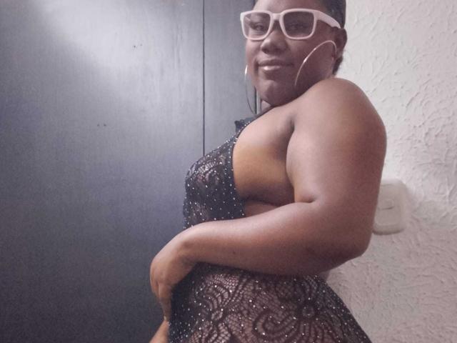 SweetyChocolatte, een donkerhuidige vrouw met een bril, draagt een doorschijnende zwarte jurk, waardoor haar grote, ronde billen en sterke arm zichtbaar zijn. Ze glimlacht lichtjes..