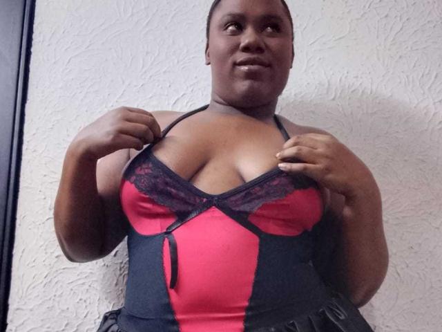 SweetyChocolatte, een mollige zwarte vrouw, staat er, gekleed in een rode en zwarte kanten lingerie-top, en trekt de bandjes naar beneden om haar grote, ronde borsten te laten zien. Ze glimlacht zelfverzekerd..