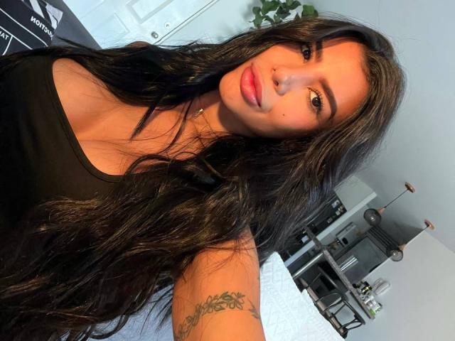 NikolRyder met lang, golvend bruin haar, roze lippen, een zwart tanktopje, een neusring, een tatoeage op de rechterarm, die naar de camera kijkt, in een lichte kamer. Borsten zichtbaar onder het topje..