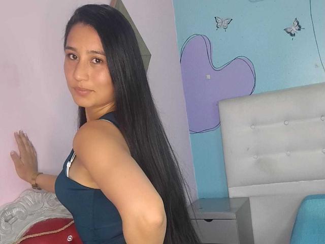Tiiffanys, com cabelos longos e pretos, vestindo uma regata preta, em pé em frente a uma parede rosa, olhando por cima do ombro, mostrando seu bumbum pequeno e seios planos..