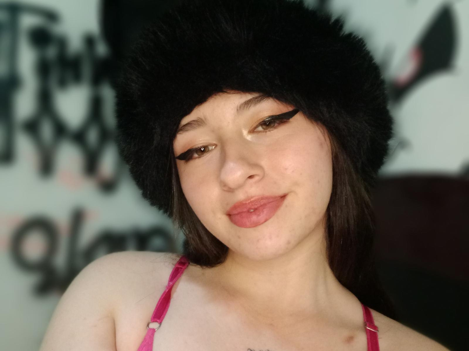 CherrySex69 with pale skin, brown eyes, and pink lipstick, wearing a black fur hat and pink bra, smiling softly. Her tits are visible, with a slight blush on her cheeks.