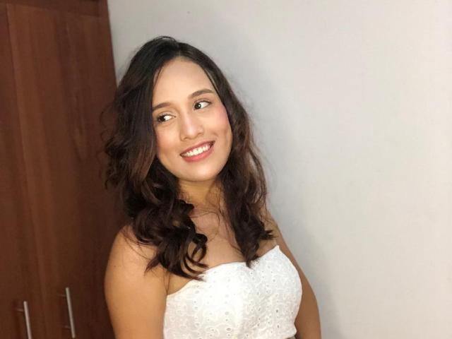 EmmaWat with wavy dark brown hair, smiling, wearing a white strapless dress, showcasing her tits, standing against a light gray wall, with a wooden cabinet on the left.