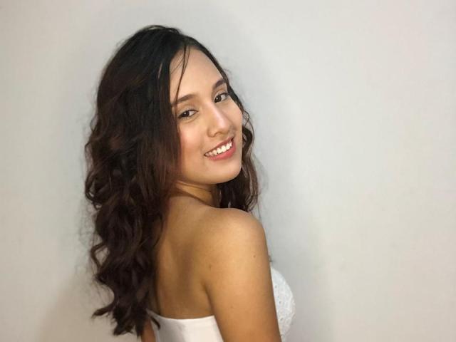 EmmaWat with long wavy brown hair, smiling at the camera, showing her bare shoulders and white strapless top, exposing her small, perky tits.