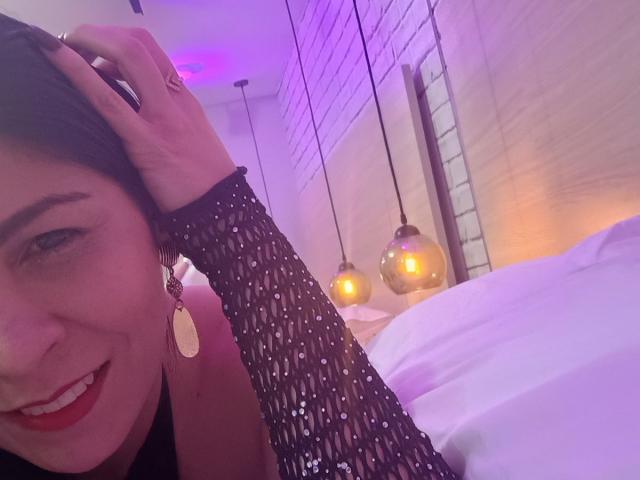El rostro de EliseBell, con lápiz labial rojo, una manga de malla negra, aretes dorados, sonriendo, con la mano en el cabello, en una cama con almohadas blancas, y luces colgantes moradas y amarillas en el fondo..