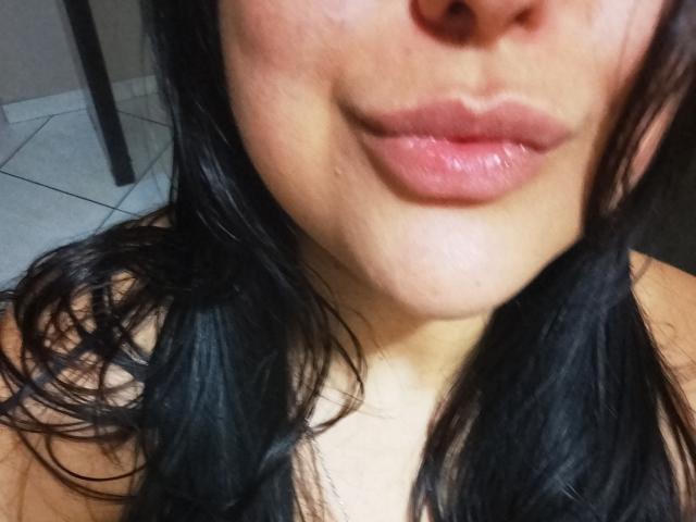Le visage de TalyaB, avec des lèvres roses et brillantes, légèrement retroussées, laissant apparaître un petit sourire. Elle a de longs cheveux noirs et ondulés, et un teint brun clair. Les seins ne sont pas visibles..