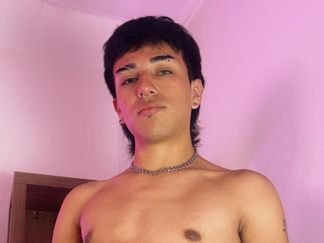 DavelSvetla has short black hair, a nose piercing, a lip piercing, and a chain necklace, with a slight smile and visible chest. Shirtless, with a tattoo on the arm.