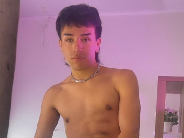 Shirtless DavelSvetla with dark hair, small nipples, chain necklace, and a nose piercing, standing in pink-lit room, looking at camera with a neutral expression.