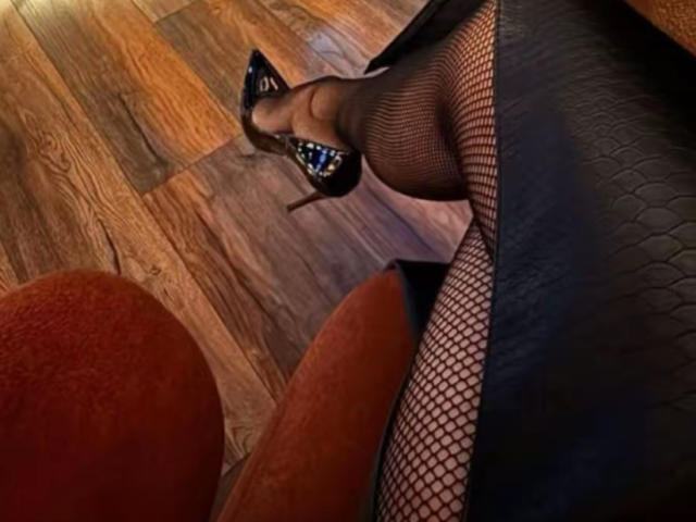 AngelinaDeleon's legs in fishnet stockings and black high heels with blue sparkles, wearing a black textured skirt, on a wooden floor with a red chair in the background.