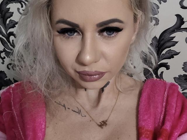 ButterflyMiss69 with platinum blonde hair, dark eyebrows, and pink lipstick, wearing a pink robe, showing a small tattoo below her chin, and a necklace with a small pendant.