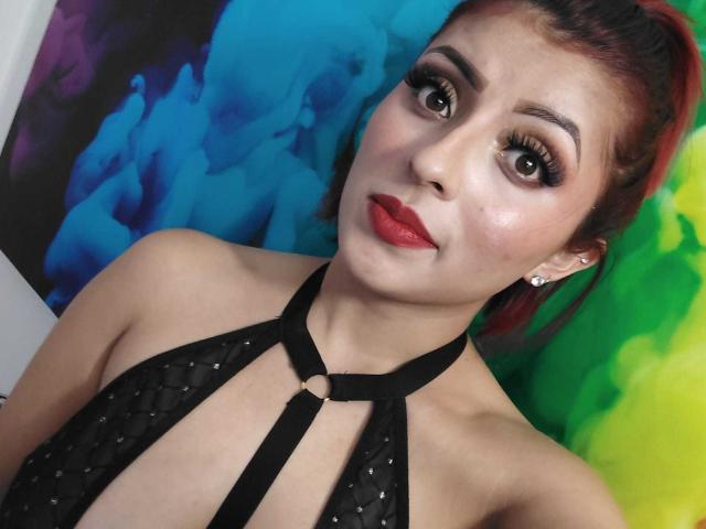 LizBonnet with red lipstick, dark eye makeup, and black halter top showing her tits, has red and brown hair, and a ring in her top strap, looking at the camera.