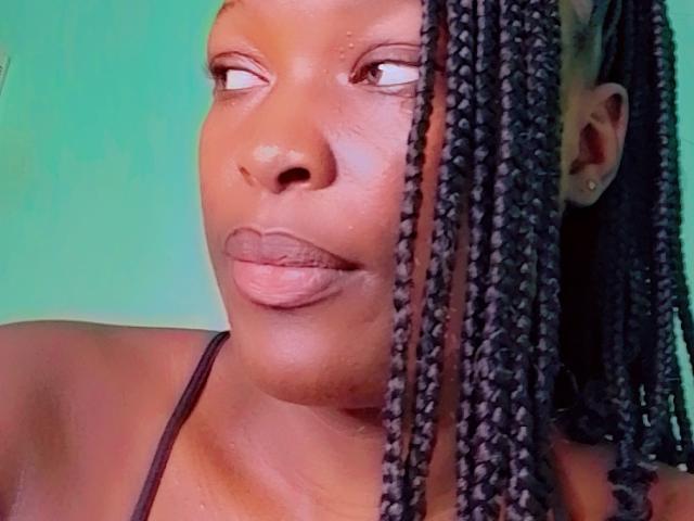 Close-up of SexyAshlee with dark brown skin. She has long, straight black braids falling over her right cheek. Her lips are slightly parted, showing a small gap in her front teeth. She wears a black halter top. Her eyes look to the right. The background is a solid green. The image has a soft, slightly blurred texture. Her face is well-lit, highlighting her smooth skin and subtle facial features.