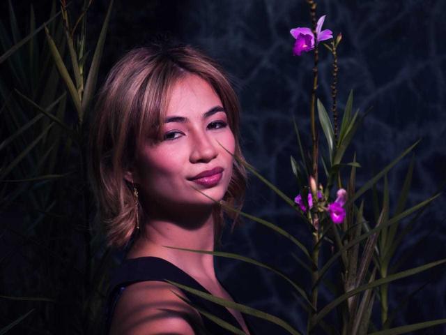MarianneBowe with short blonde hair, pink eyeshadow, and black top, smiling at camera, surrounded by green leaves and pink orchids, dark background, soft lighting, subtle makeup.