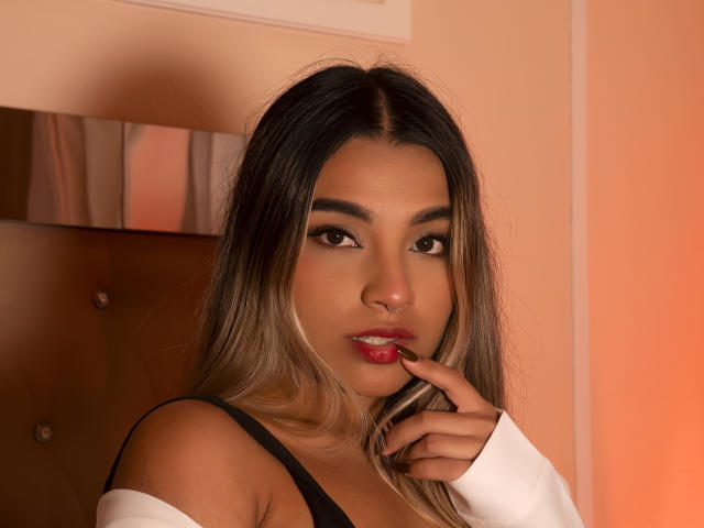 AnneBull with long dark blonde hair, brown eyes, red lipstick, nose ring, white off-shoulder top, black strap, finger near mouth, soft orange light, sexy, attractive, subtle smile.