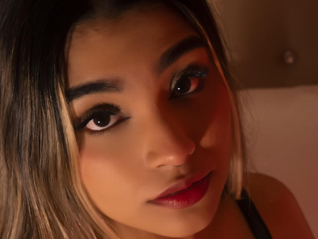 AnneBull's face, brown eyes, black eyeliner, subtle makeup, red lipstick, straight blonde and black hair, soft brown skin, slight smile, warm light, close-up, gentle expression, sexy look.