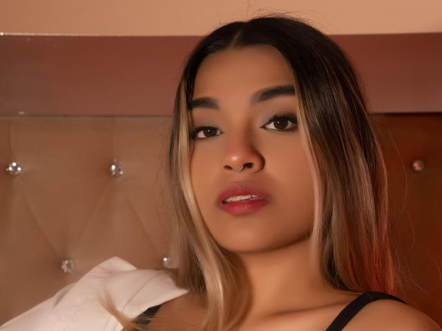 AnneBull has brown eyes, light brown skin, and long blonde-streaked hair. She's looking at the camera with pink lipstick, black strap, and a white pillow in the background.