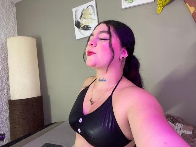 MelodyBlossom has pink-tinted skin, black hair, and is wearing a black bra with buttons, eyes closed, red lipstick, and a tattoo on her neck. Her tits are visible.