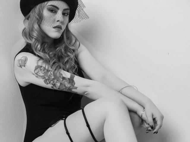 RoseanneFaded with wavy blonde hair, black hat, and mesh veil, wearing a black dress and garter, sitting with crossed legs, showing a detailed tattoo on her right arm.