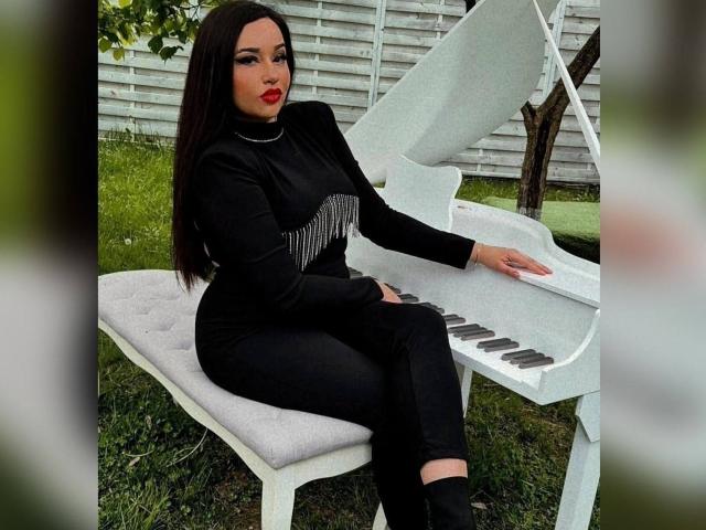 RachelleDom met donker haar, rode lippenstift, een zwarte jurk en broek, zittend op een witte pianobank, spelend op een witte vleugel, handen op de toetsen, billen op de zitting, gras buiten..