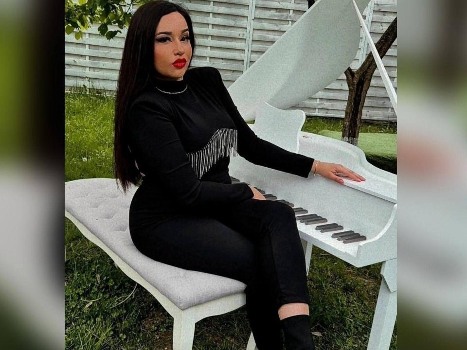 RachelleDom, con cabello oscuro, lápiz labial rojo, vestido negro y pantalones, sentada en un banco blanco para piano, tocando un piano de cola blanco, con las manos sobre las teclas, el trasero en el asiento, y césped afuera..