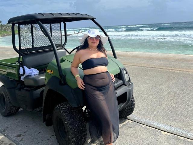 LilyRousee stands next to a green utility vehicle with black mesh sides. She's wearing a white sunhat, black strapless top, and sheer black skirt. Her dark hair is loose, and she has a gold bracelet on her left wrist. The vehicle has a small white cloth on the seat. The vehicle is parked on a concrete surface near the ocean with waves in the background. The sky is mostly clear with some clouds. She leans slightly to the right with her right hand on the vehicle.