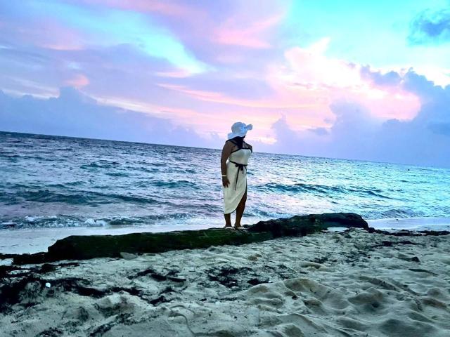 LilyRousee stands on a rocky beach, wearing a white headscarf and dress, holding a baby, looking at the ocean under a pink and blue sunset sky.