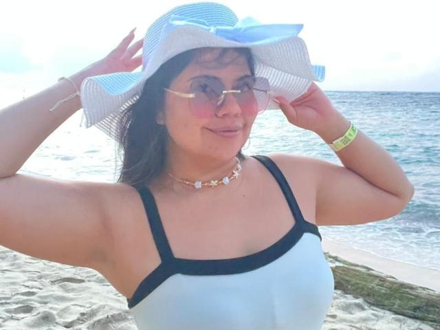 LilyRousee, medium-skinned woman with wavy dark hair, wears a light blue strapless top with black trim, a white sun hat with a blue ribbon, pink-tinted sunglasses, and a star-shaped choker. She holds the hat's brim with both hands, smiling slightly. Her right wrist has a yellow-green bracelet. The beach and ocean are in the background. Bright sunlight enhances the scene. She has a relaxed, casual appearance.