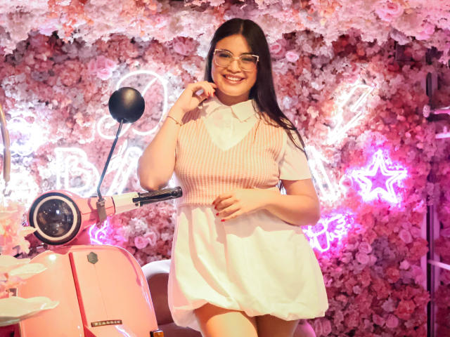 CamilaSofiia stands in a pink, floral-themed room. She has long dark hair, glasses, and a bright smile. She wears a pink and white checkered dress with short sleeves. Her right hand is near her mouth, and her left rests on her thigh. A pink scooter with a black handle is to her left. Neon pink star and lettering are in the background. The image is colorful and playful.