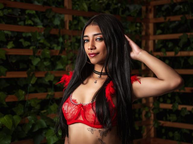 LoreneLash met lang zwart haar, rode kant BH met veren, zwarte choker, hand in haar, kleine tatoeage onder de borst, lachend, donkere lipstick, gemiddelde borsten, bruine huid, groene ogen..