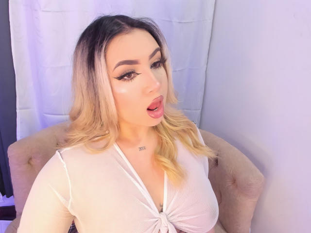 MelissaLust with blonde hair, open mouth, and heavy eyeliner, wearing a white top that shows her tits, sitting on a beige chair, white curtain in the background.