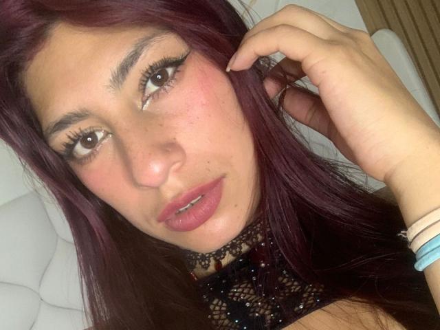 AlisonCold with purple hair, brown eyes, and pink lips, wearing a black lace choker, touching her ear, looking at the camera, with a ring and blue bracelet on.